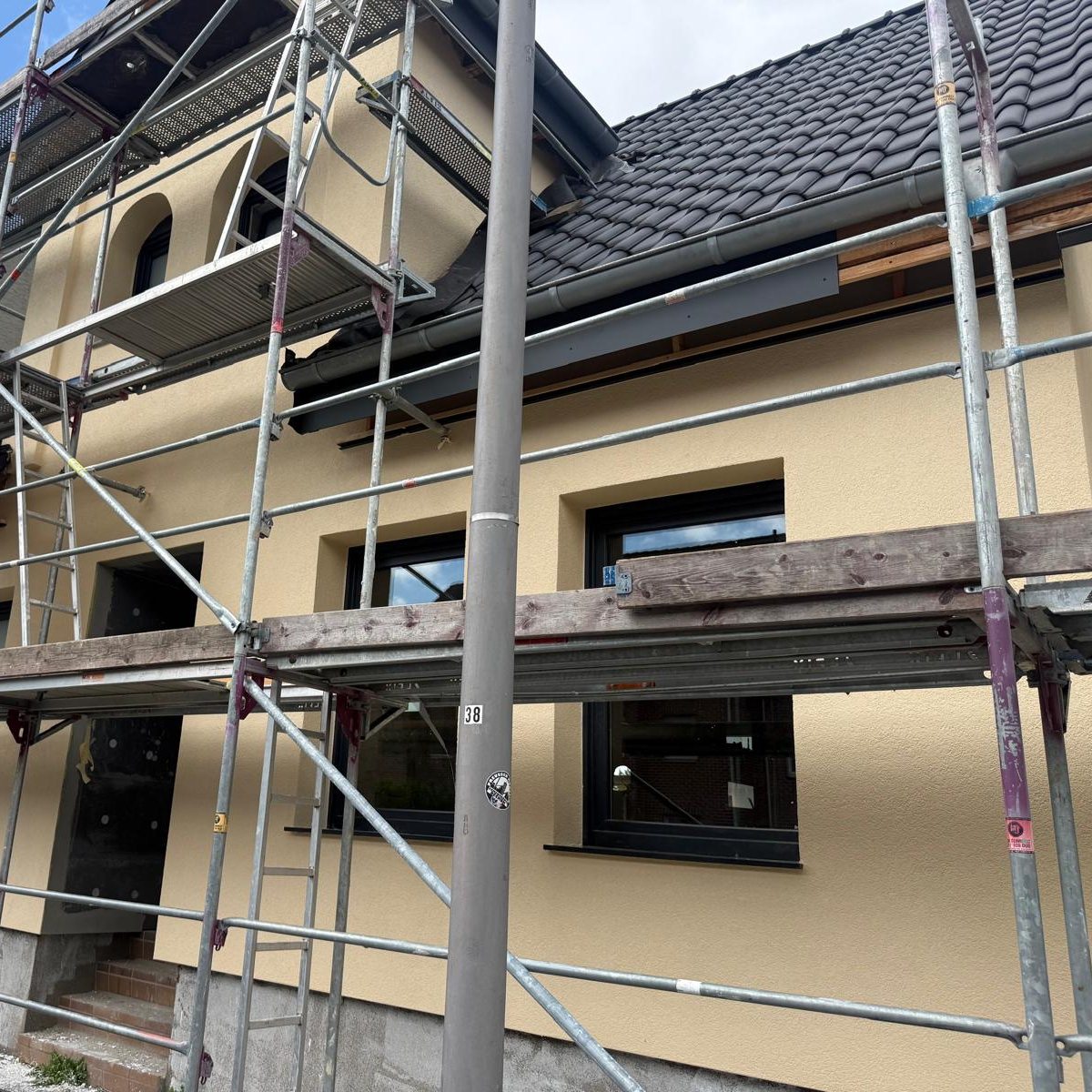 Gelb verputztes Haus mit dunklem Dach und neuen Fenstern hinter einem Baugerüst