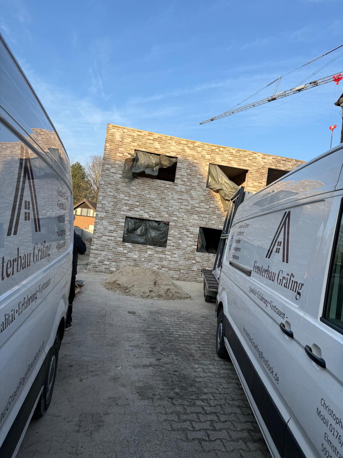 Blick zwischen zwei Firmenwagen von Fensterbau Gräfing auf einen Klinkerrohbau