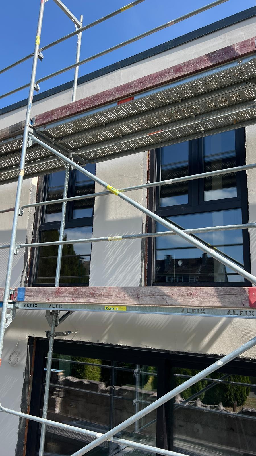 Dunkel gerahmte Fenster in heller Fassade hinter Gerüstbohlen