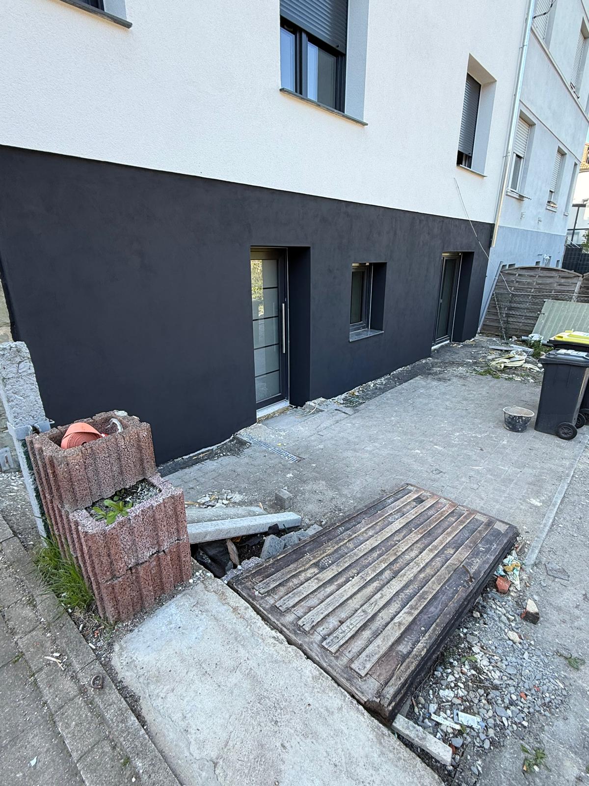 Hausecke mit dunklem Sockel, Nebentür und Fenstern, davor ein Hof mit Bauresten