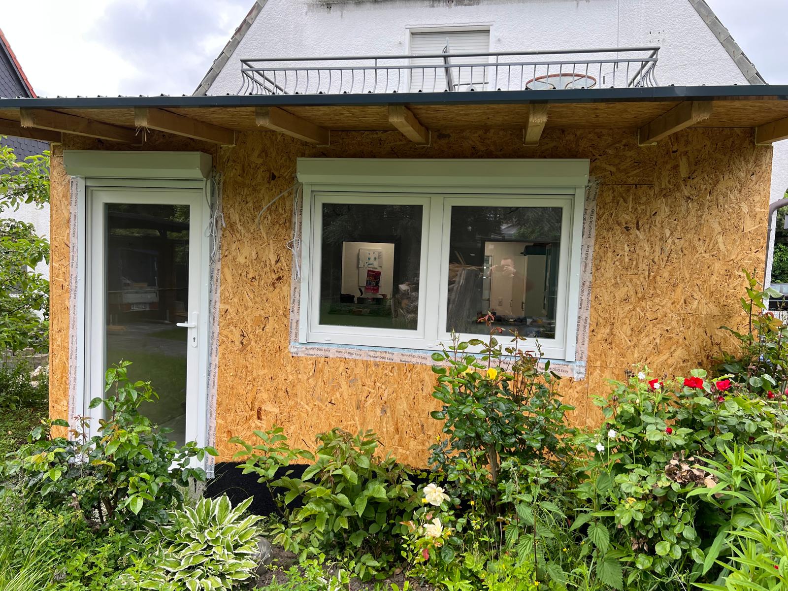 Anbau mit noch unverkleideten Holzwänden, einem weißen zweiflügeligen Fenster und einer weißen Glastür, davor ein Rosenbeet