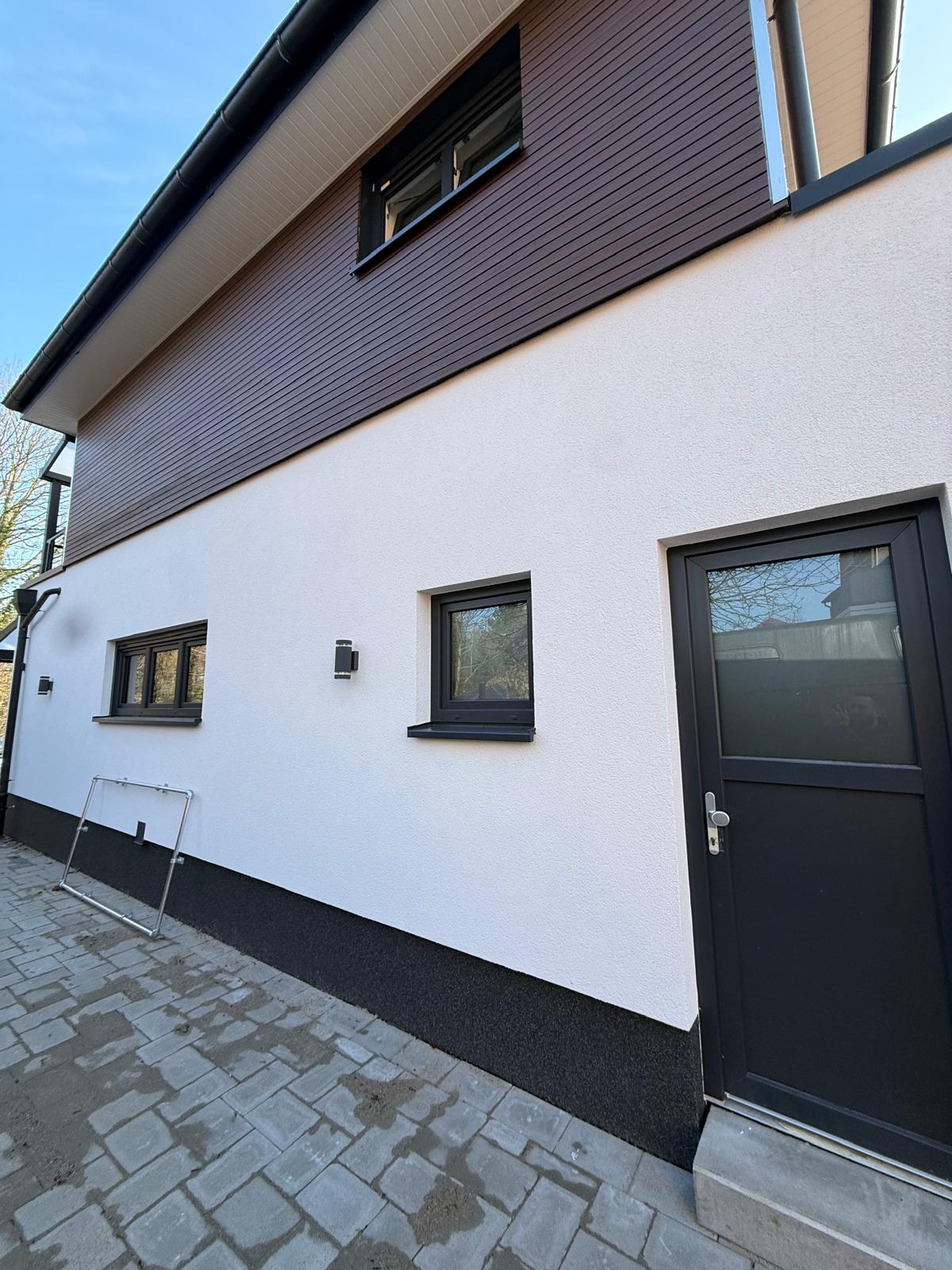 Anthrazitfarbene Nebeneingangstür mit Glasausschnitt in einer weißen Fassade, darüber eine Holzverkleidung