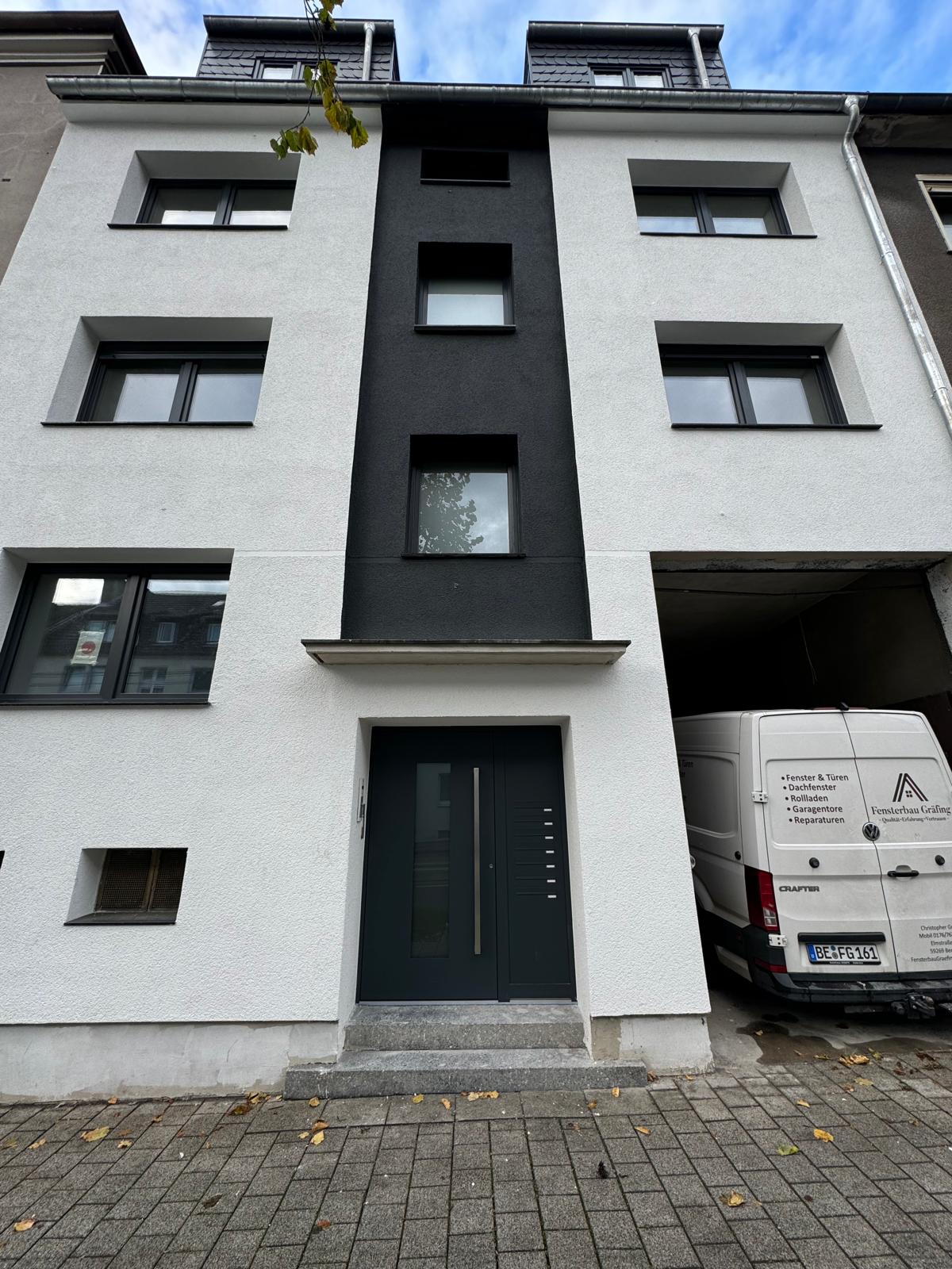 Weiß-dunkle Fassade eines Mehrfamilienhauses mit anthrazitfarbener Haustür, daneben der Firmenwagen in der Durchfahrt