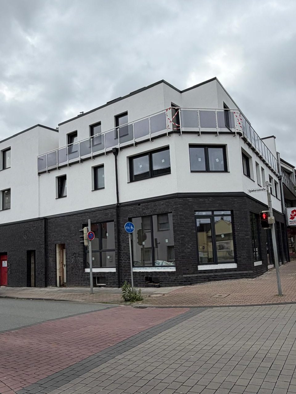 Mehrgeschossiges Eckgebäude mit dunklem Klinkersockel, weißer Fassade und Dachterrasse unter bewölktem Himmel