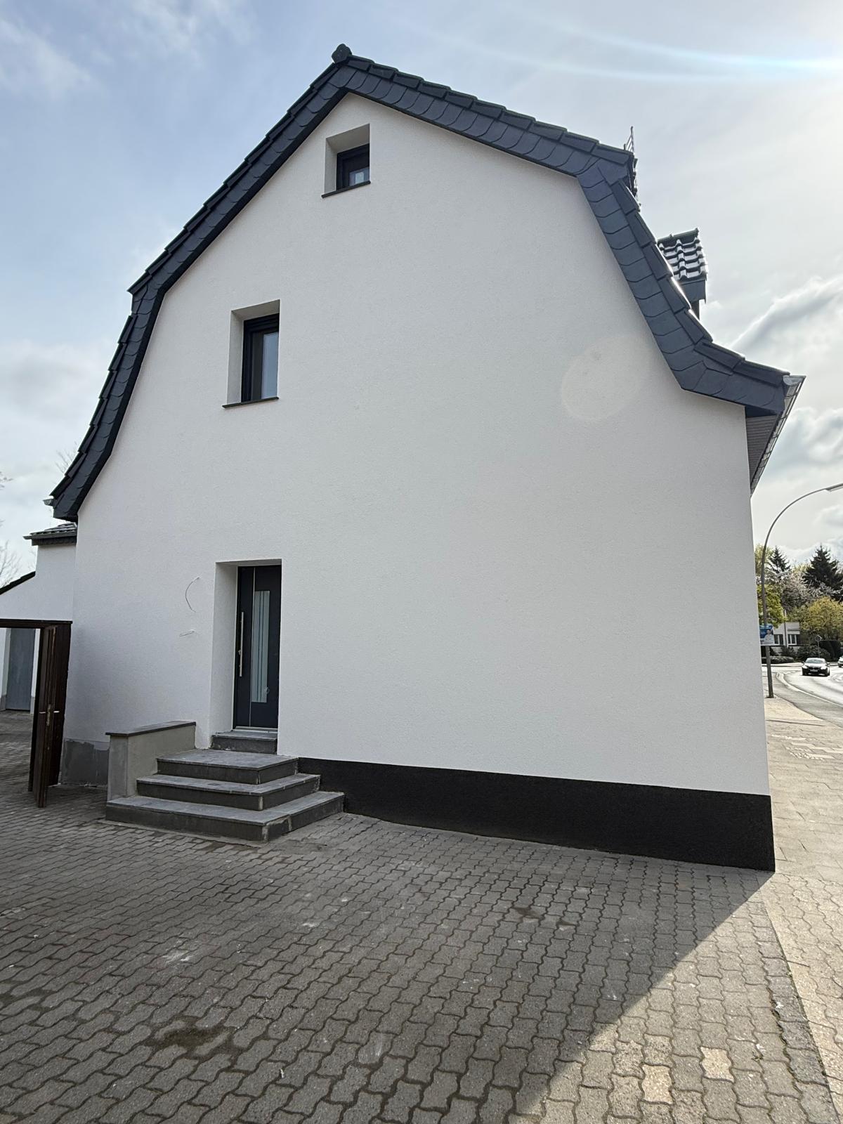Weiße Giebelseite eines Hauses mit dunklem Sockel, kleinen Fenstern und einer Tür mit Treppe