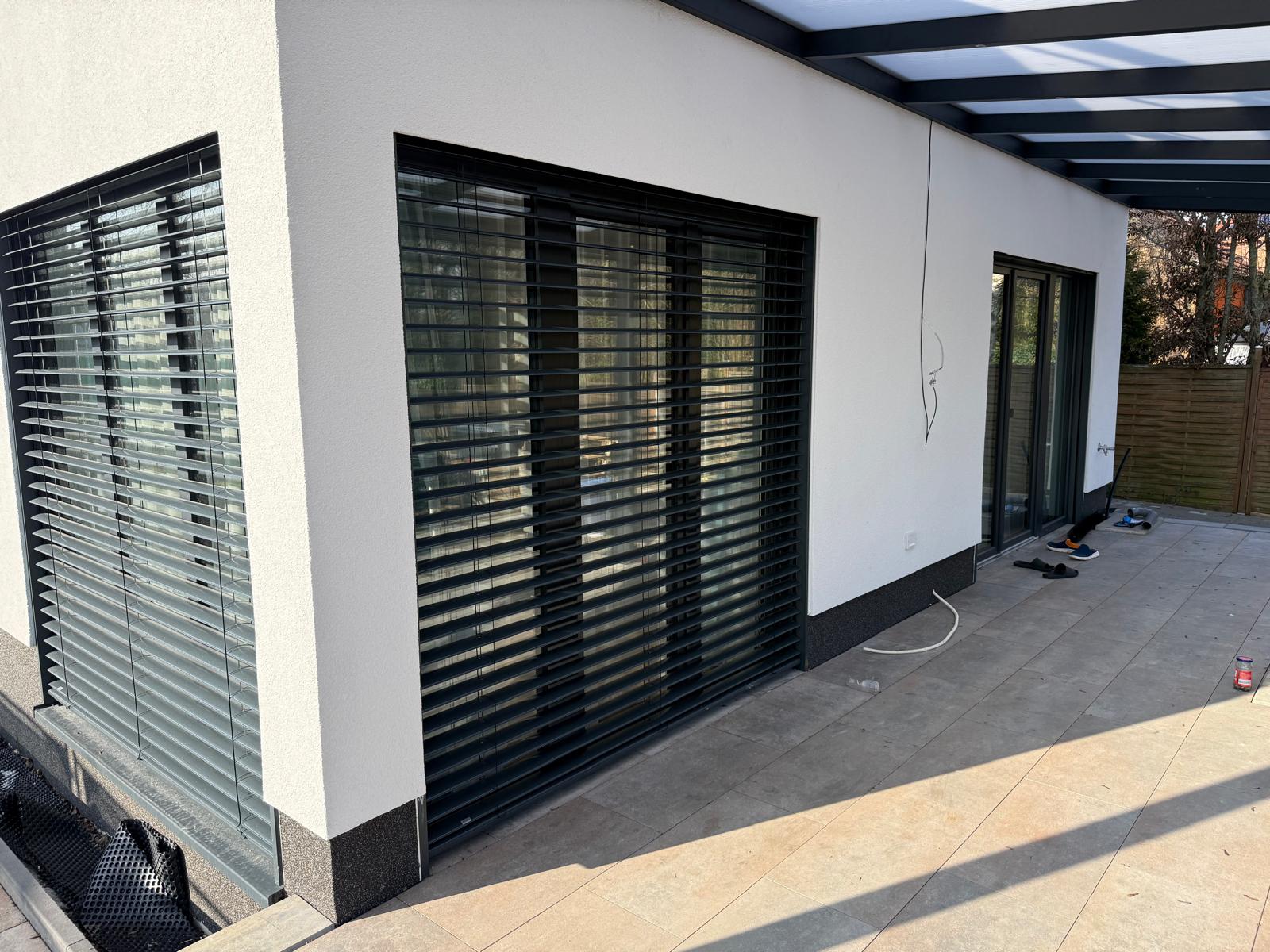 Weiße Hauswand mit bodentiefen Fenstern hinter dunklen Außenjalousien an einer Terrasse