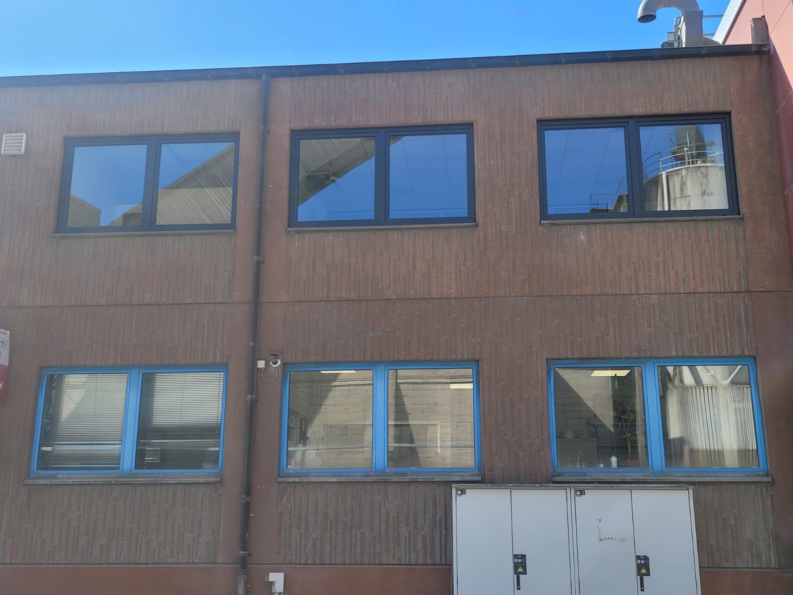 Zweigeschossiger Gewerbebau mit rotbrauner Fassade, dunklen Fensterrahmen oben und blauen unten