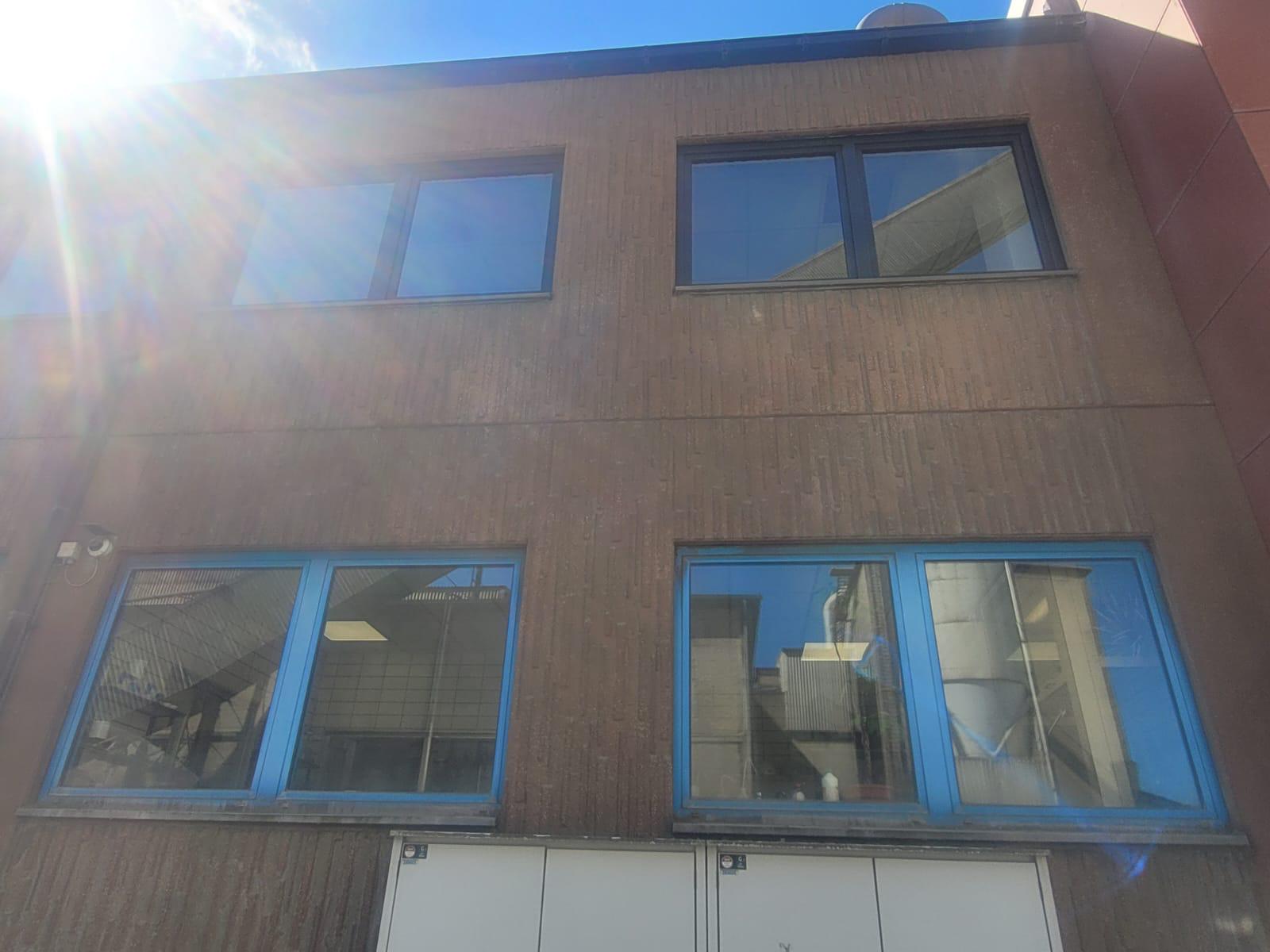 Rotbraune Gewerbefassade mit dunklen Fenstern oben und blau gerahmten Fenstern unten, darüber die Sonne