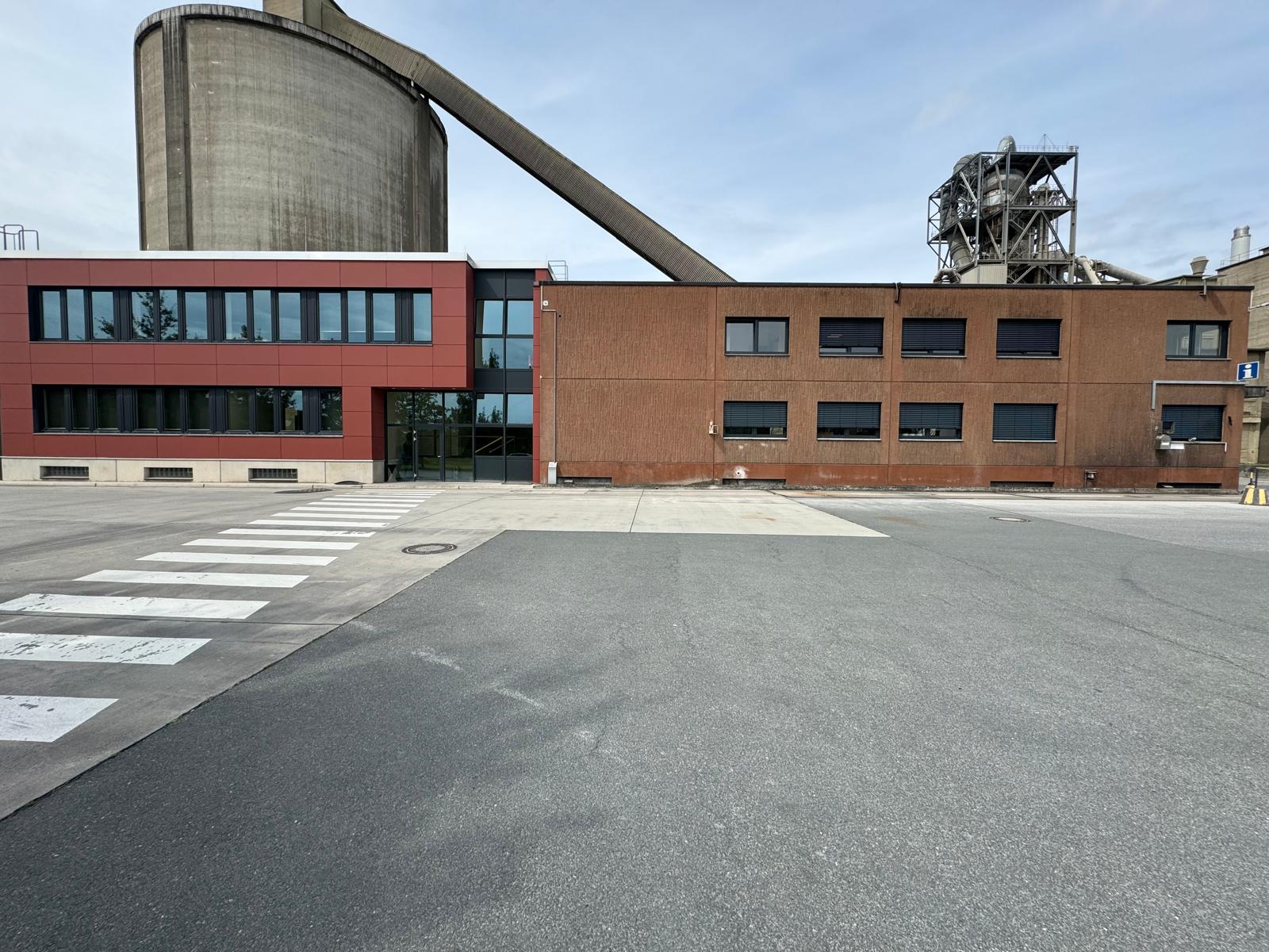 Flacher Gewerbebau mit dunklen Fenstern neben einem roten Bürogebäude, dahinter ein Silo