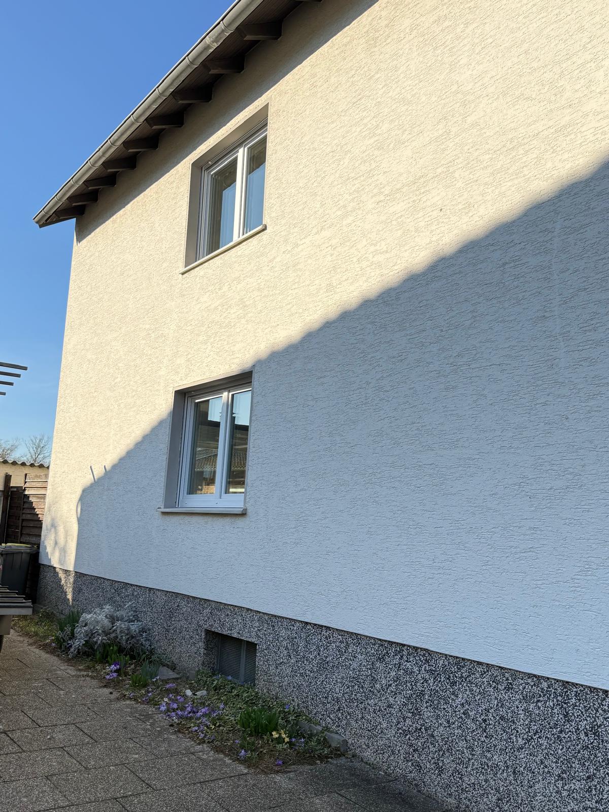 Seitenwand eines hell verputzten Hauses mit zwei weißen Fenstern unter blauem Himmel