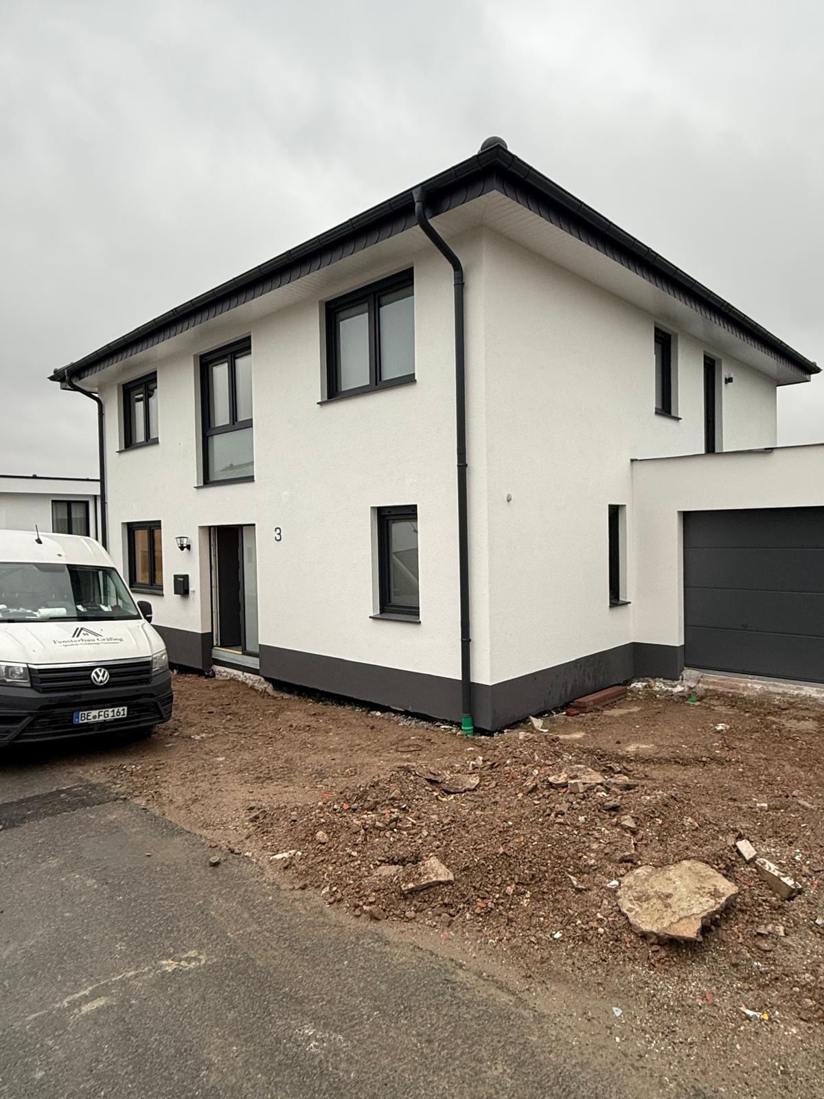 Weißer Neubau mit dunklem Sockel, anthrazitfarbenen Fenstern und dunklem Garagentor, davor der Firmenwagen