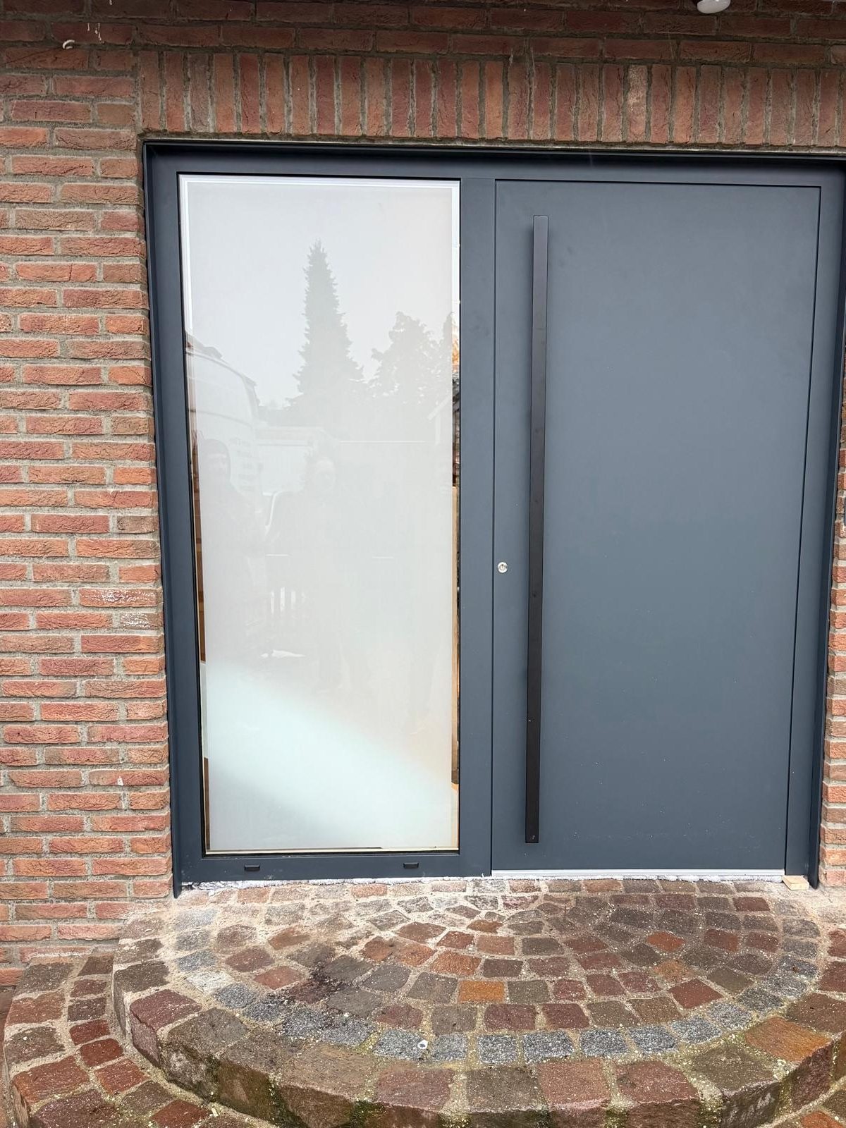 Anthrazitfarbene Haustür mit mattem Glas-Seitenteil in einer Backsteinfassade, davor runde gepflasterte Stufen